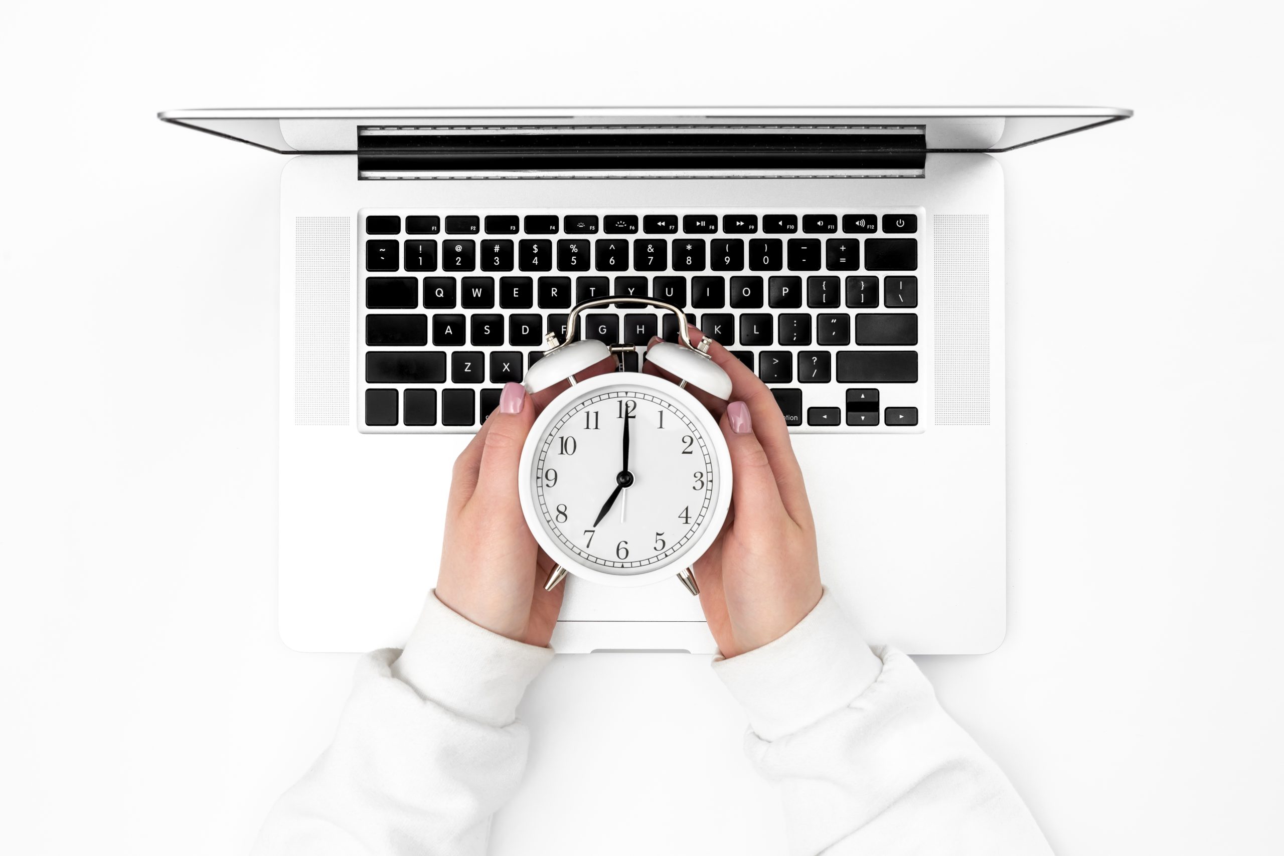 Alarm clock in female hands and a laptop on a white background, top view. Minimalistic background, the concept of work in the office, mornings at work, schedule and deadline.